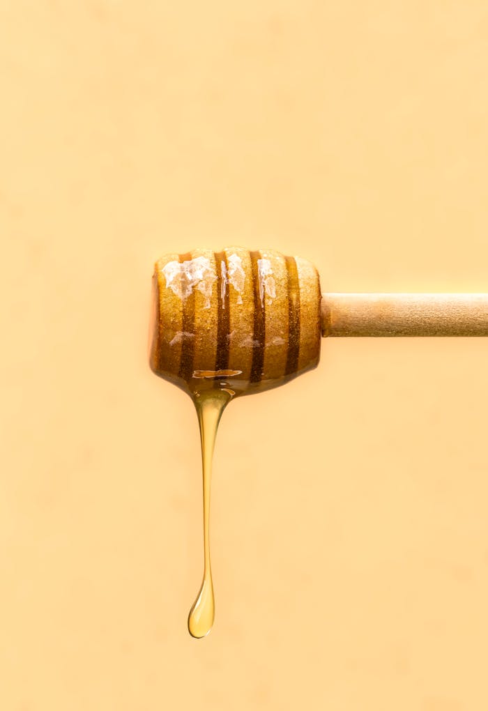 Close-up of honey drizzling from a wooden dipper against a soft yellow background.